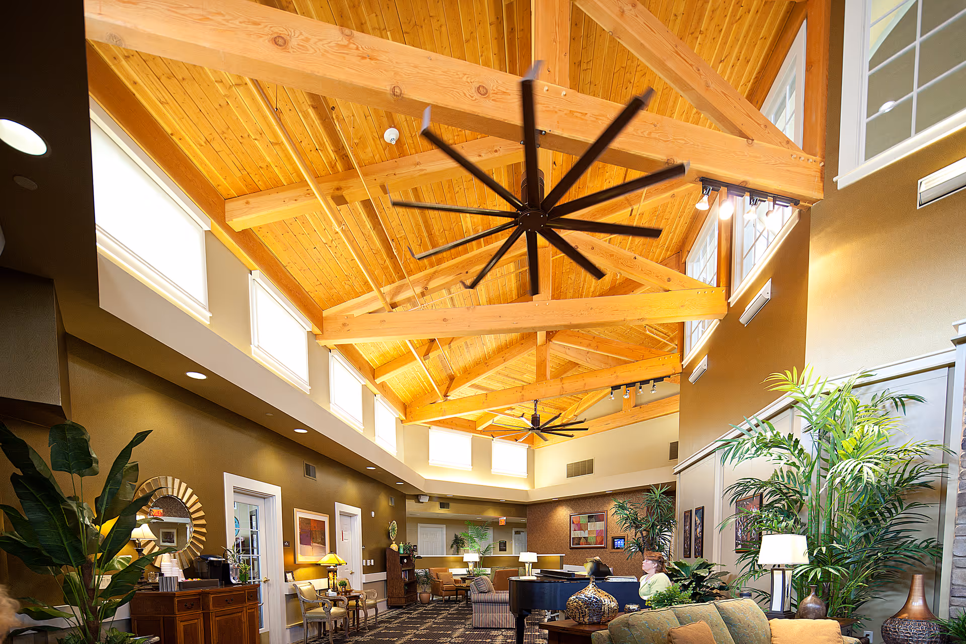 A spacious and well-lit living room area in a retirement community with high wooden vaulted ceilings, large ceiling fans, multiple windows near the ceiling, comfortable seating including sofas and armchairs, indoor plants, lamps, and a grand piano. The room has a warm and inviting atmosphere with a mix of modern and rustic decor.