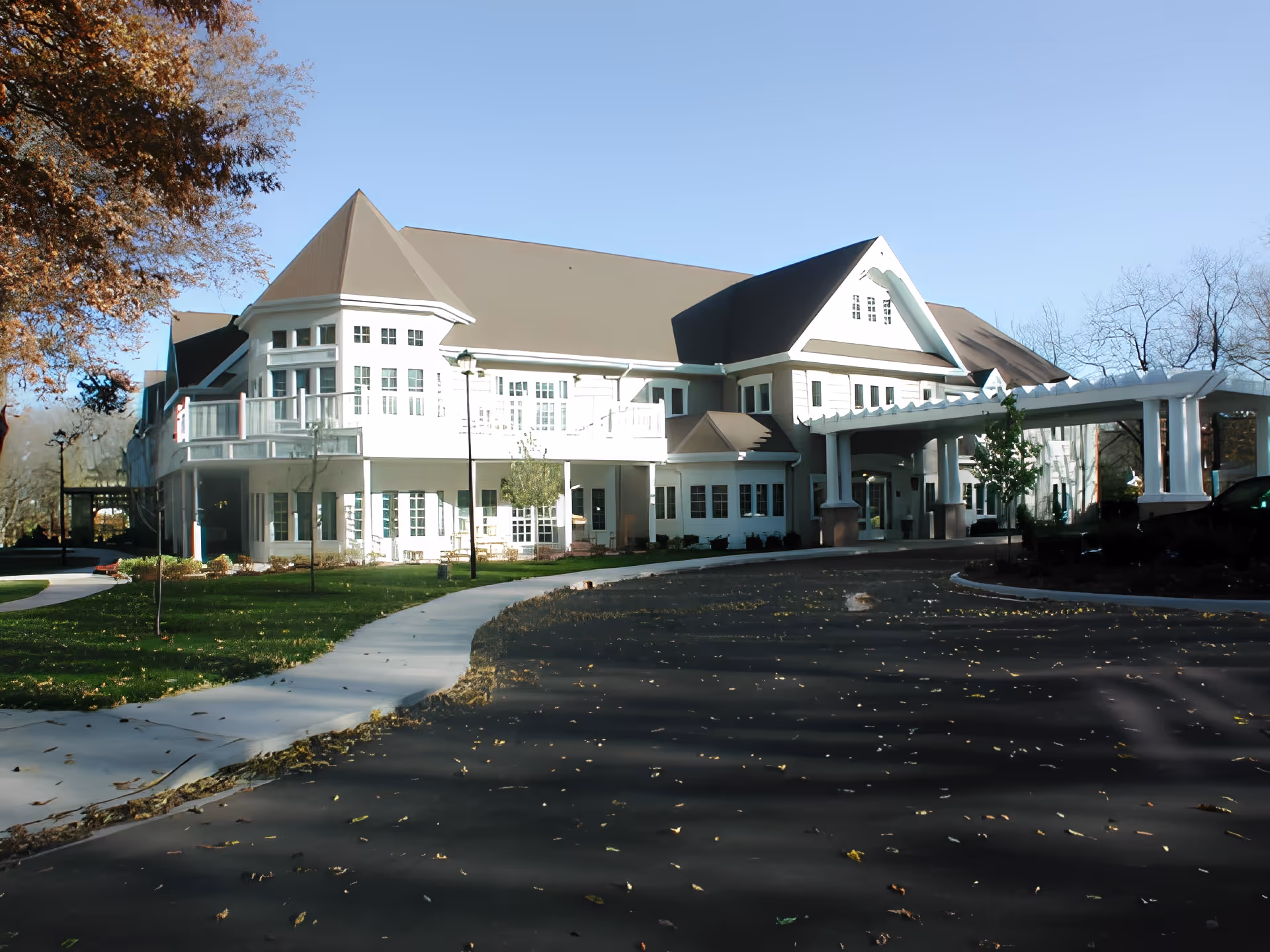 Exterior view of a large senior living facility building with white walls and a brown roof, surrounded by a paved driveway and green lawn with trees. The building features multiple windows and a covered entrance with white pillars.