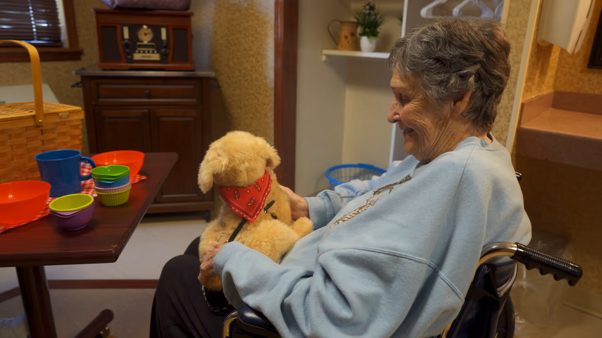 An elderly woman in a wheelchair smiling and holding a stuffed dog wearing a red bandana in a cozy room with a table set with colorful bowls and cups.