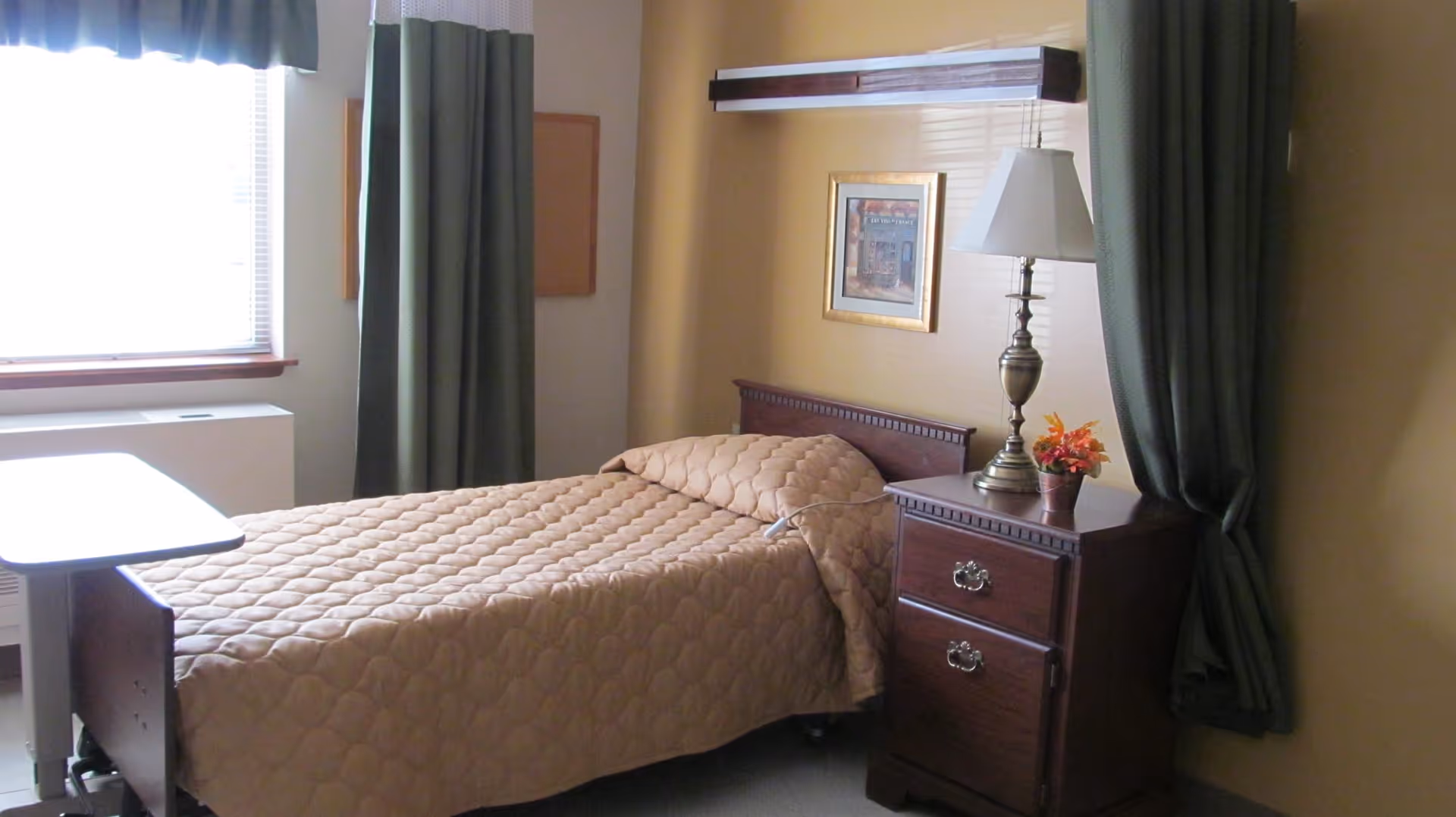 A single bed with a beige quilted bedspread in a room with tan walls. Next to the bed is a wooden nightstand with two drawers, a brass lamp with a white lampshade, and a small flower pot with orange flowers. There is a window with green curtains and a valance, and a framed picture hanging on the wall above the bed.