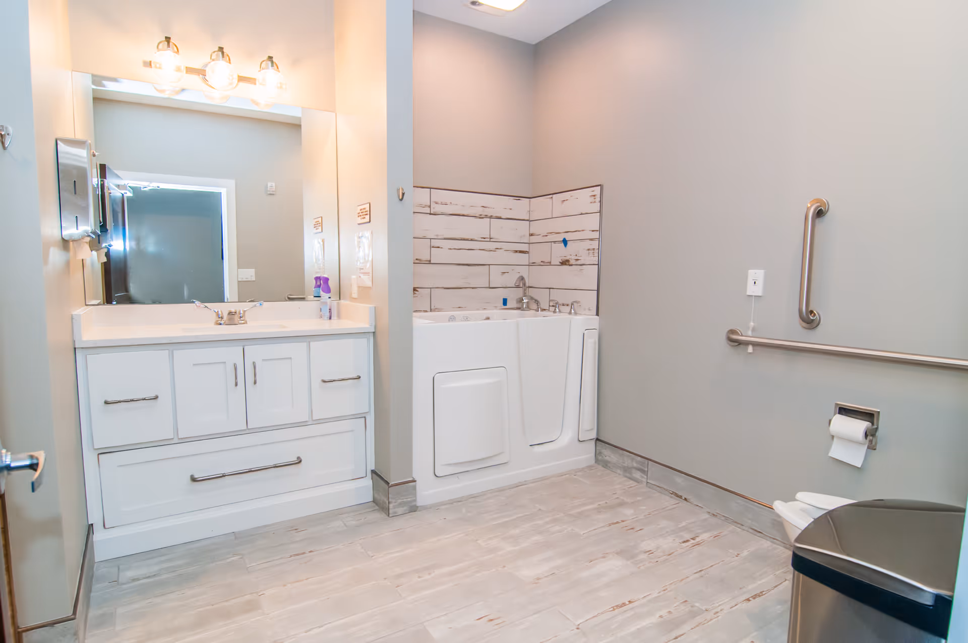 A spacious, accessible bathroom with a white vanity and sink, a large mirror with three light fixtures above it, a walk-in bathtub with a door, grab bars on the walls, a toilet with a toilet paper holder, and a trash can. The walls are painted light gray and the floor has light-colored wood-like tiles.