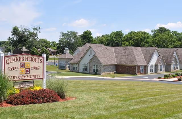 Exterior view of Ohio Living Quaker Heights care community building with a large sign in the foreground reading 'Quaker Heights Care Community' and a well-maintained lawn and landscaping under a partly cloudy sky.