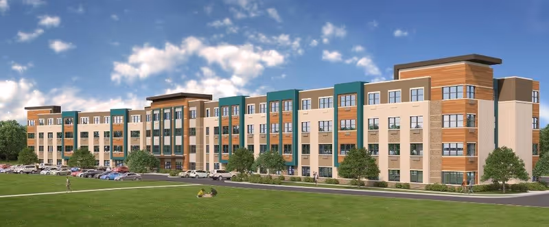 Exterior view of a modern four-story senior living facility building with a mix of beige, green, and wood panel accents. There are several windows, a parking area with cars, and a green lawn with a few people walking and sitting. The sky is partly cloudy with blue patches.