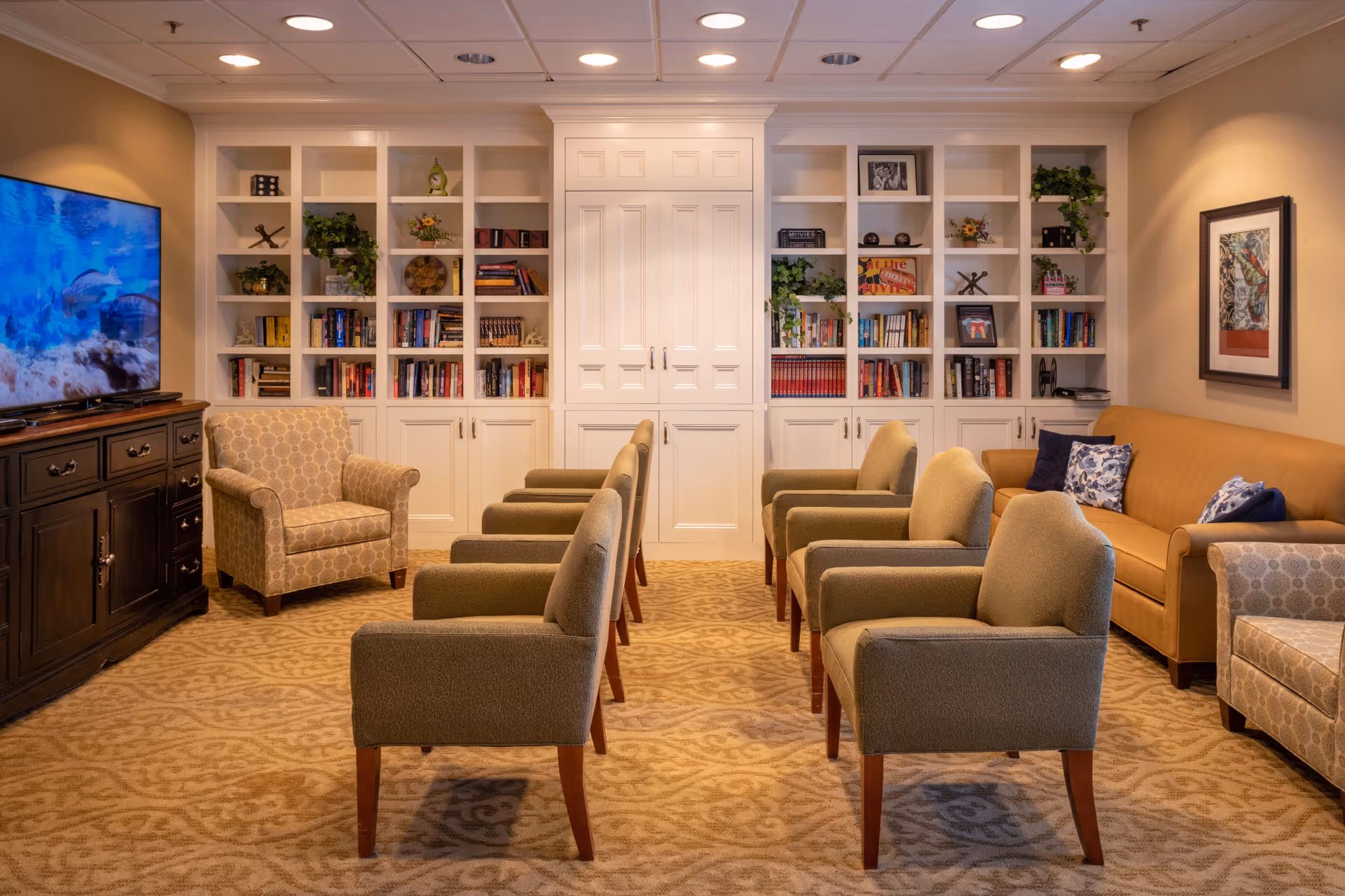 A cozy living room with six beige armchairs arranged in two rows facing a large flat-screen TV on a dark wooden cabinet. The room features a patterned carpet, a tan sofa with decorative pillows, two patterned armchairs, built-in white bookshelves filled with books and decorative items, and warm lighting.