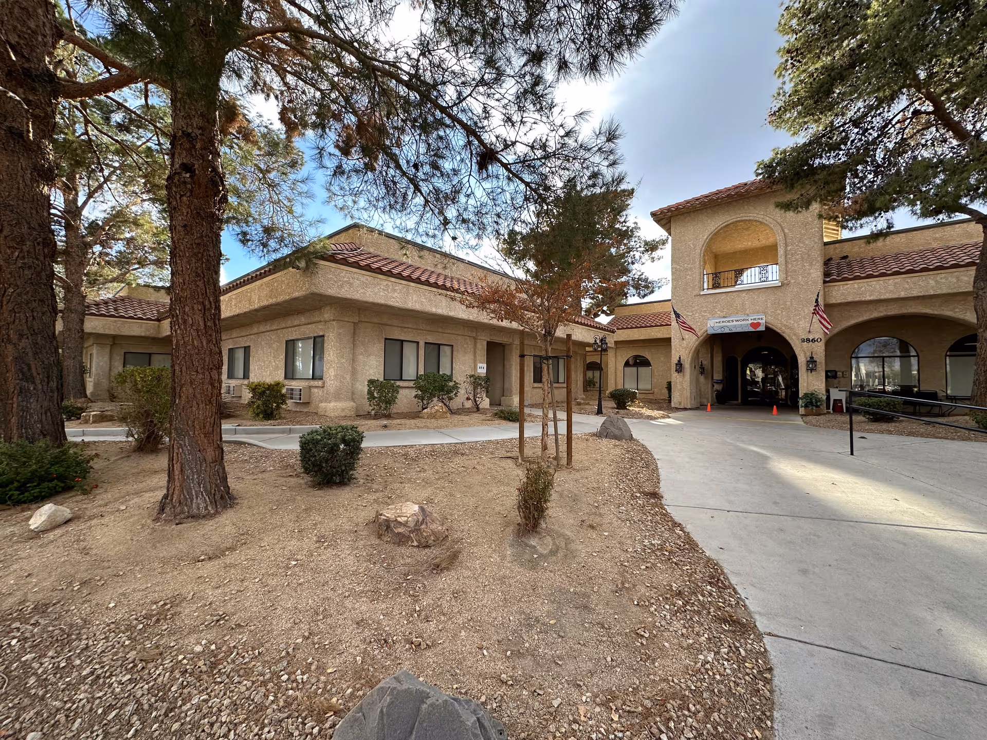 Exterior view of Mission Pines Nursing & Rehabilitation Center showing a beige stucco building with red tile roofing, surrounded by trees and shrubs. A paved walkway leads to an arched entrance with two American flags and a banner that reads 'HEROES WORK HERE'.