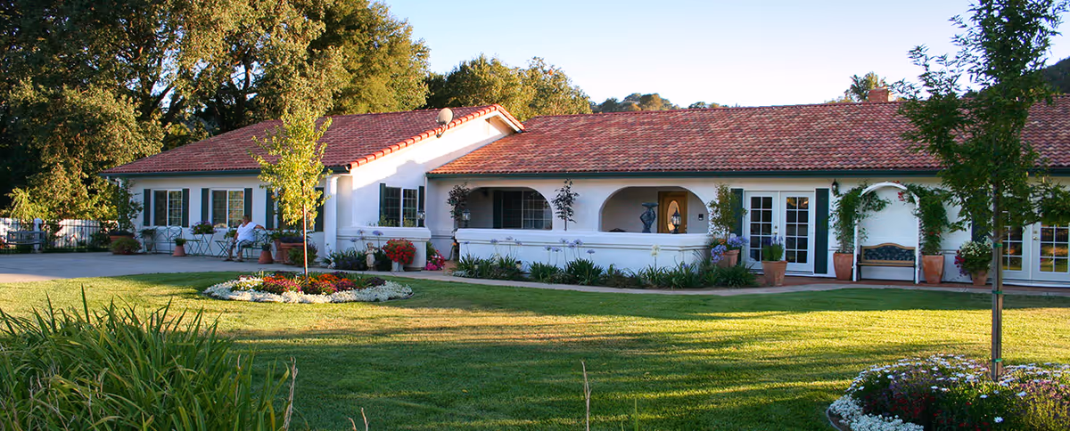 A single-story building with white walls and a red tile roof, surrounded by a well-maintained lawn and flower beds. There are several potted plants and small trees near the building, and a person sitting on a bench on the left side. The scene is bright and sunny with large trees in the background.