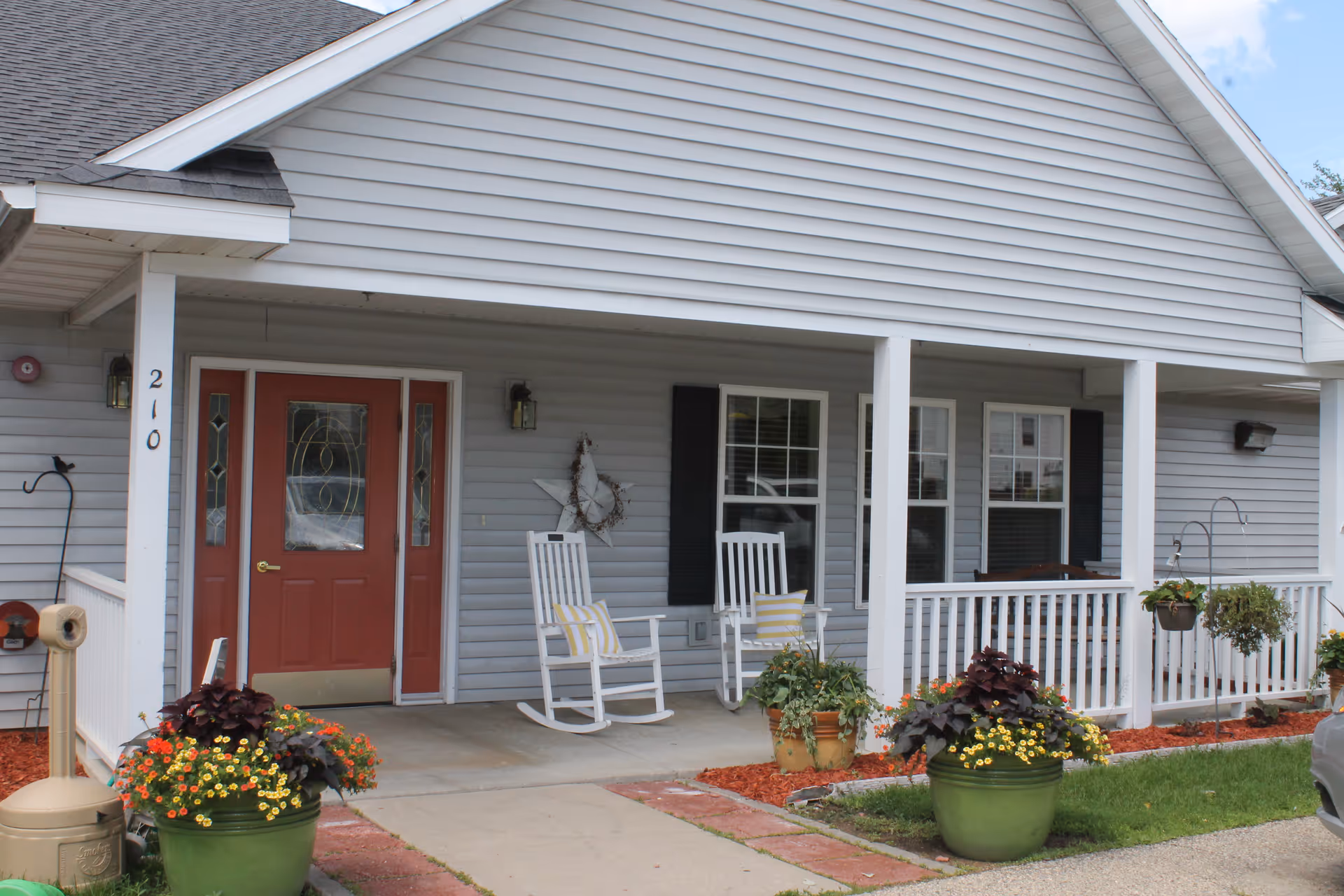 Front porch of a residential-style building with gray siding and a red door. Two white rocking chairs with yellow and white striped cushions are placed on the porch. There are potted plants with colorful flowers and greenery along the walkway and porch. The building number 210 is visible on a white column.