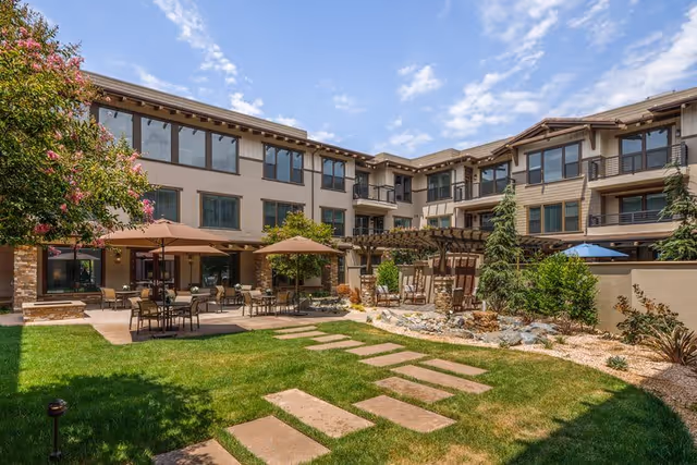 Outdoor courtyard area of a senior living facility with green grass, stone pathway, patio tables with umbrellas, trees, and a three-story building surrounding the courtyard under a partly cloudy sky.