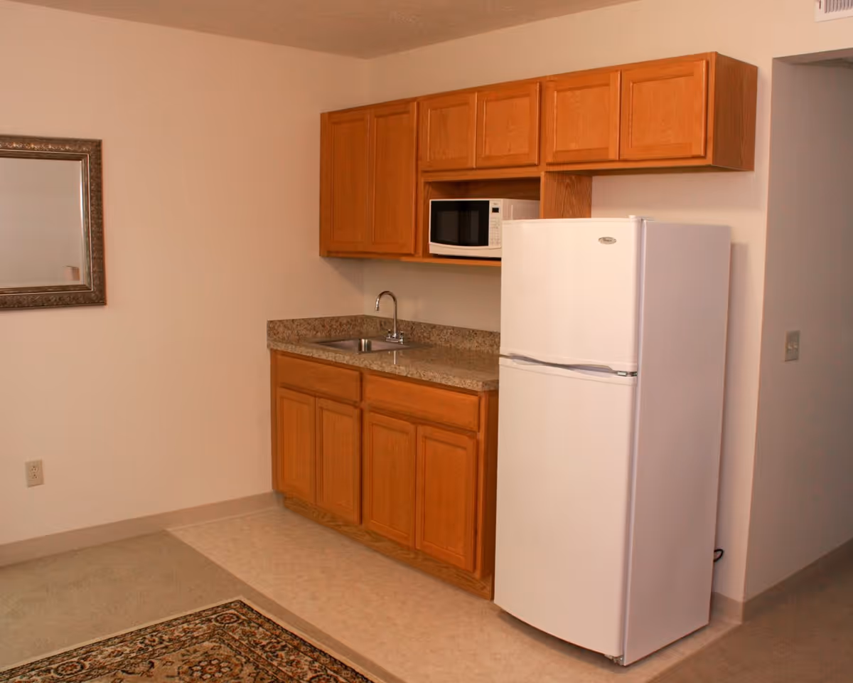 Small kitchenette area with wooden cabinets, a granite countertop with a sink, a white refrigerator, and a microwave oven. There is a framed mirror on the adjacent wall and a patterned rug partially visible on the floor.