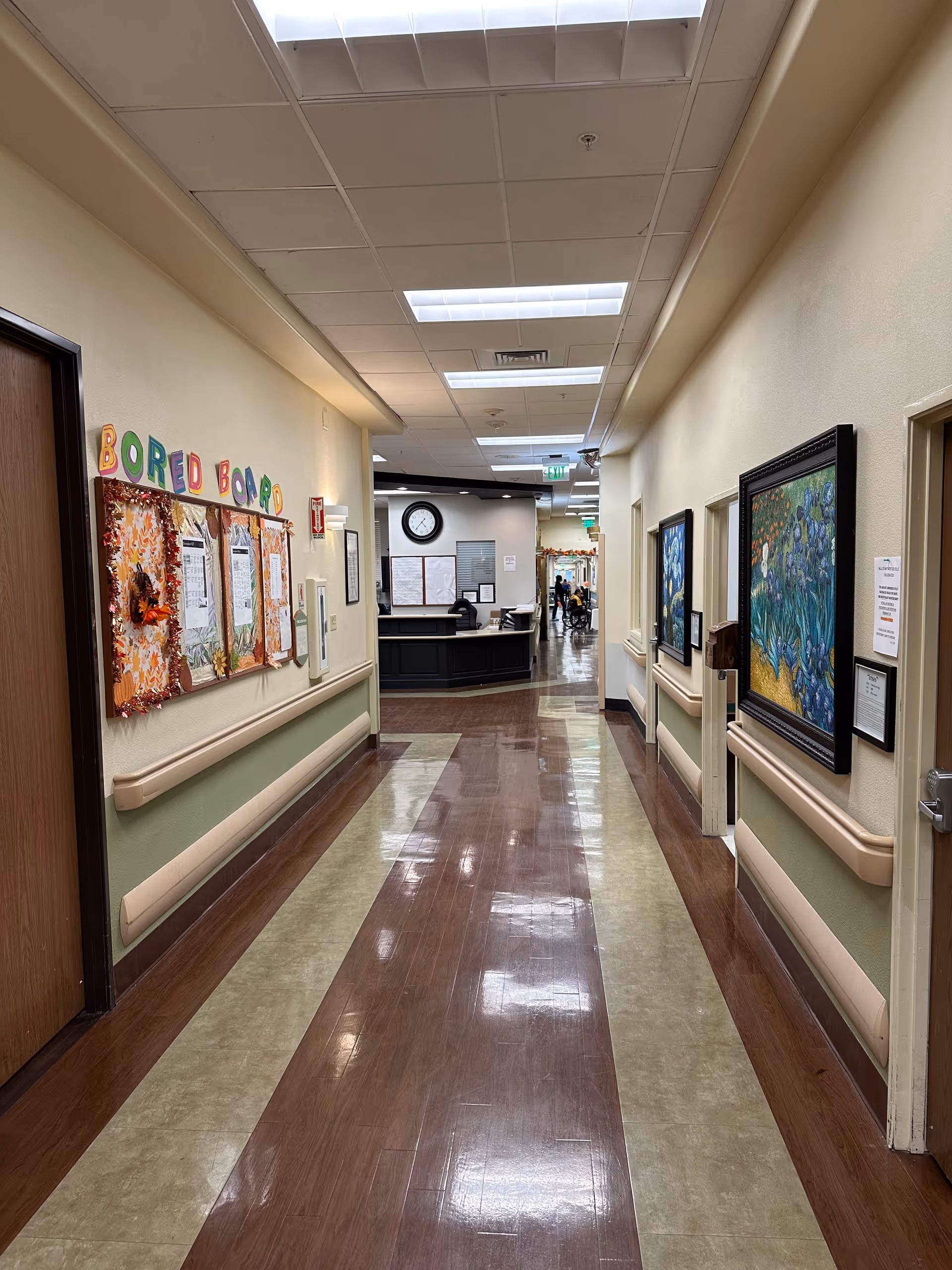 A clean, well-lit hallway in a nursing and rehabilitation center with polished floors, handrails on both sides, bulletin boards with colorful decorations on the left wall, framed artwork on the right wall, and a reception desk visible at the end of the corridor.