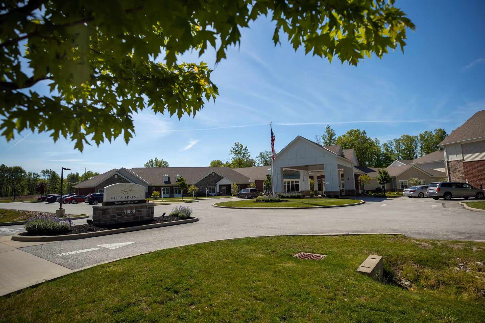 Exterior view of Vista Springs Quail Highlands facility on a sunny day with a clear blue sky. The building has a covered entrance with an American flag, surrounded by a circular driveway and parked cars. Green grass and trees frame the scene.