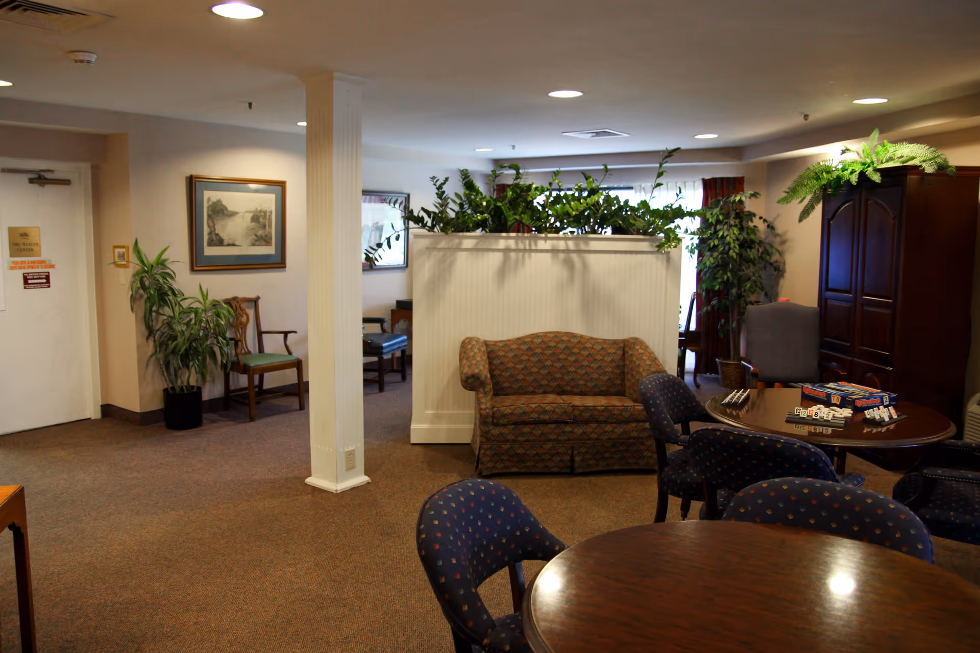 A cozy common area in a senior living facility featuring a patterned loveseat, several upholstered chairs around wooden tables, a cabinet with plants on top, and framed artwork on the walls. There are also some board games on one of the tables and various potted plants adding greenery to the space.