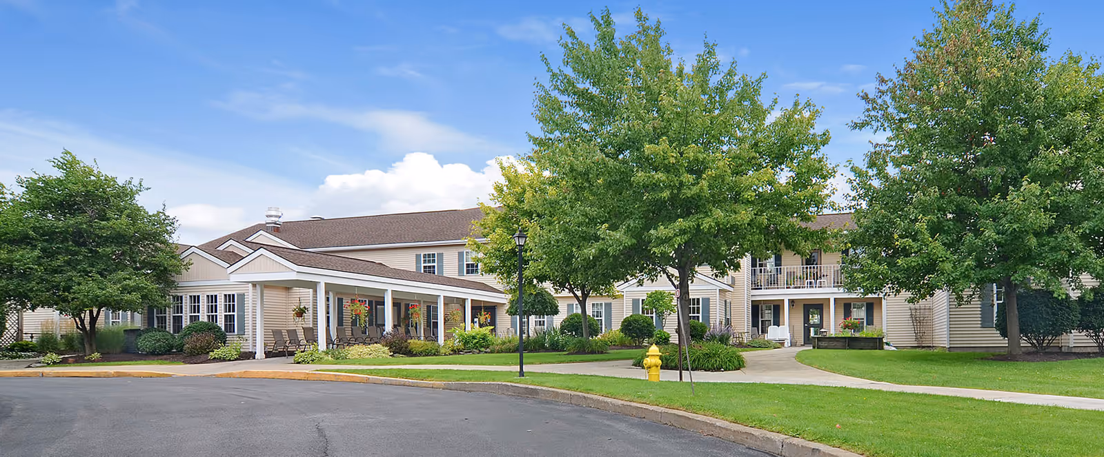 Exterior view of Brookdale Summerfield senior living facility showing a two-story building with beige siding, a covered porch with seating, well-maintained green lawns, trees, and a curved driveway under a partly cloudy blue sky.