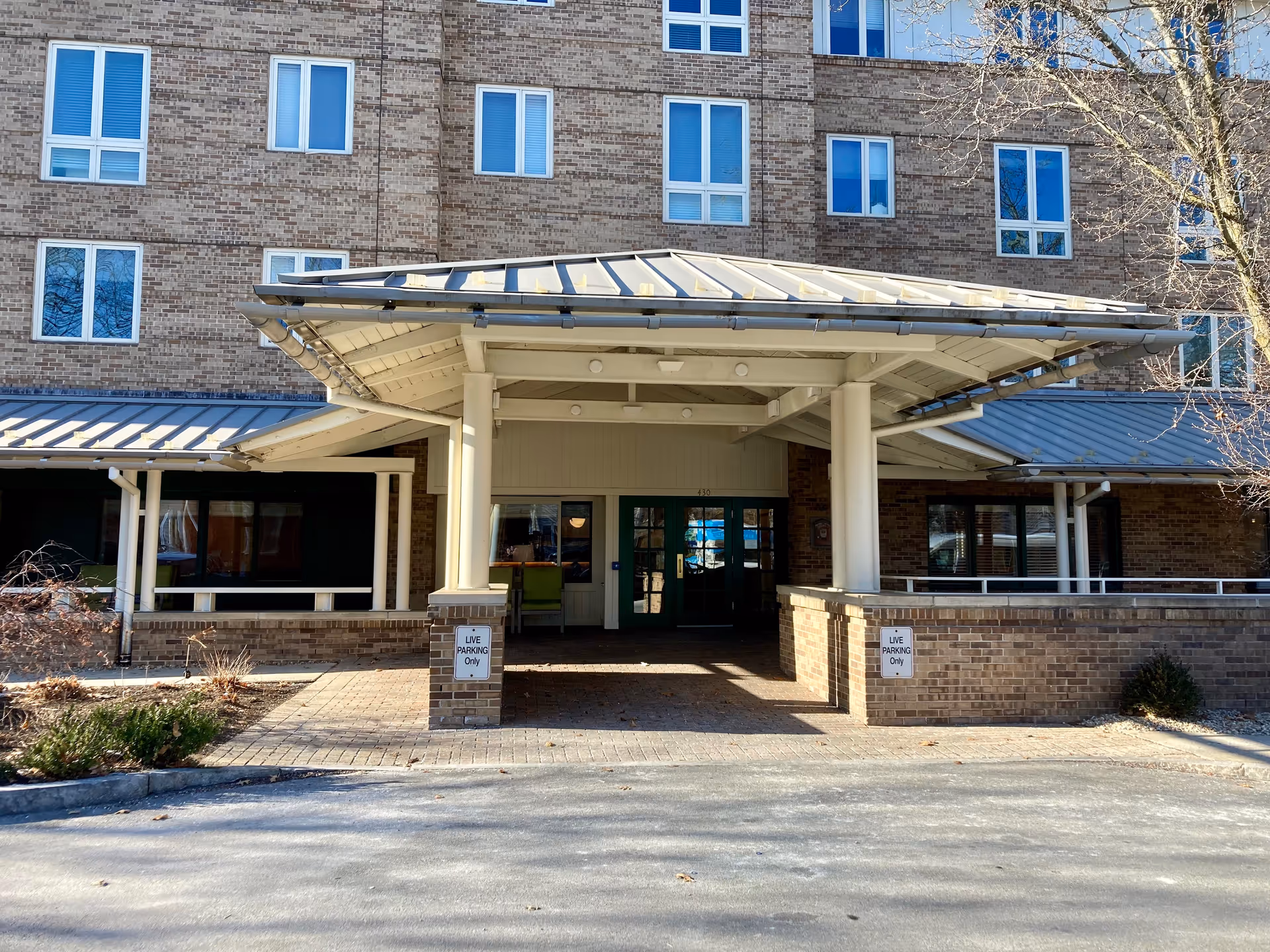 Front entrance of a multi-story brick building with a covered driveway and green double doors. There are signs on brick pillars that read 'LIVE PARKING Only'. Leafless trees and some shrubs are visible around the entrance.