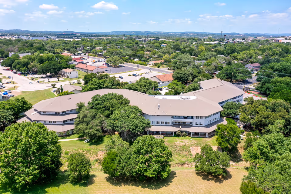 Aerial view of a large senior living facility building surrounded by many trees and greenery, with a parking lot and roads visible in the background under a partly cloudy sky.