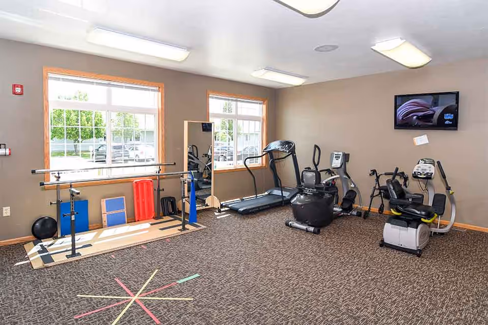 A well-lit exercise room with large windows, featuring various fitness equipment including a treadmill, stationary bikes, parallel bars, and exercise mats. A wall-mounted TV is visible on the right side.