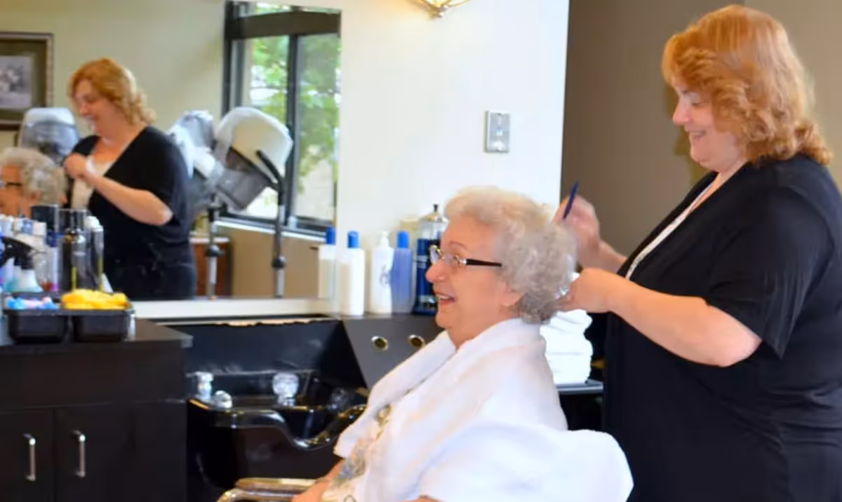 An elderly woman with white hair and glasses is sitting in a salon chair, smiling while a hairstylist with curly red hair is working on her hair. The salon has a large mirror, hair care products on the counter, and hair dryers in the background.