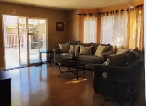 A living room with a large gray sofa and a matching armchair, both adorned with striped and solid cushions. There is a round coffee table in front of the sofa with a small flower arrangement on it. The room has wooden flooring and large windows covered with sheer curtains, allowing natural light to fill the space. A glass sliding door leads to an outdoor area.