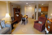 A cozy living room area in a senior living facility with a red recliner chair, a gray sofa with patterned cushions, a wooden dining table with chairs, a wooden hutch displaying dishes, and a kitchen area in the background.