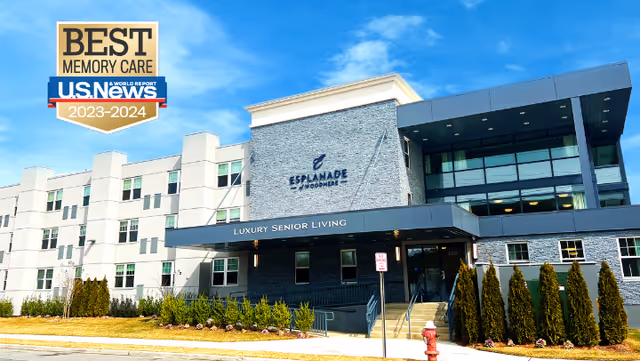 Exterior view of Esplanade of Woodmere, a luxury senior living facility with a modern three-story building featuring large windows, a covered entrance, and landscaped greenery in front. A badge in the top left corner indicates it was awarded Best Memory Care by U.S. News for 2023-2024.