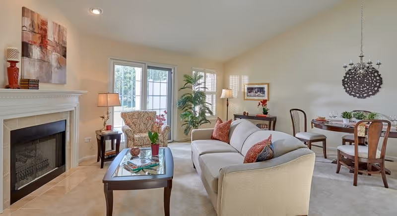 A bright and cozy living room with a beige sofa adorned with colorful pillows, a patterned armchair, a glass coffee table with books and a plant, a fireplace with decorative items on the mantel, a side table with a lamp, a potted plant near the window, and a round dining table with four chairs and a hanging light fixture in the background.