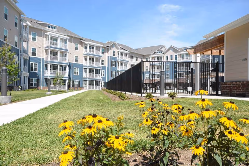 Outdoor view of The Crossings Retirement Community showing a multi-story residential building with balconies, a grassy lawn, a paved walkway, and yellow flowers in the foreground under a clear blue sky.