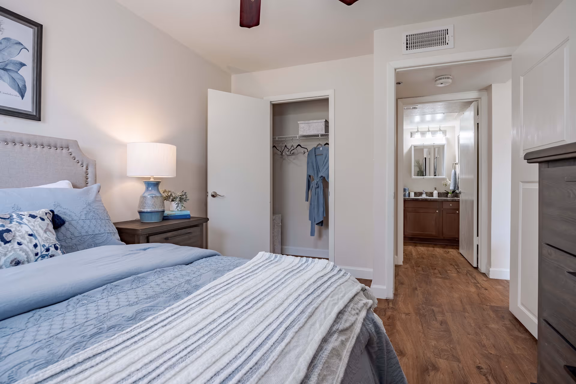 A cozy bedroom with a neatly made bed featuring blue and white bedding. A nightstand with a lamp and decorative items is beside the bed. The closet door is open, showing a hanging blue robe and some hangers. The room has wooden flooring and leads to a bathroom with a vanity and mirror visible through an open door.