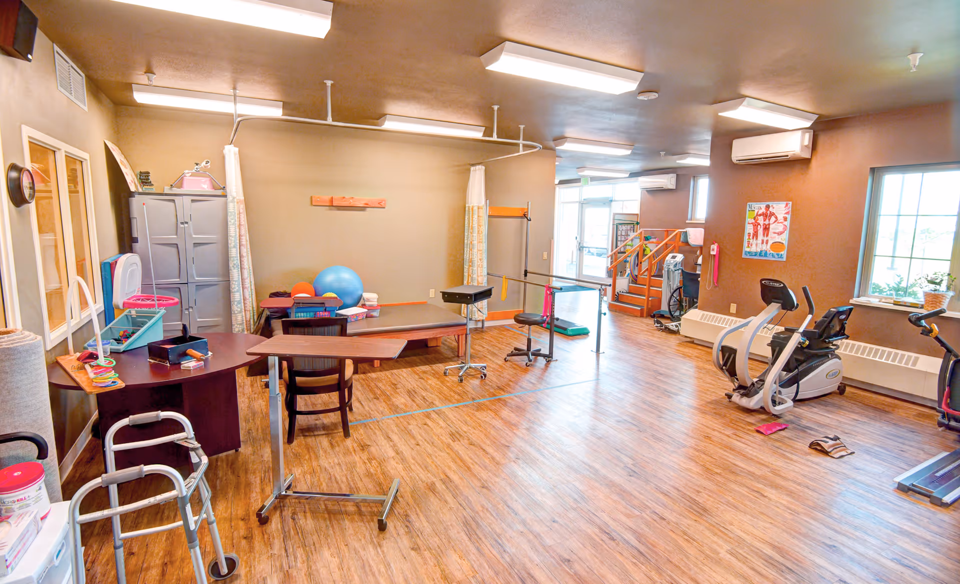 A spacious physical therapy or rehabilitation room with wooden flooring, exercise equipment including stationary bikes, therapy tables, a walker, exercise balls, and parallel bars. The room has beige walls, large windows letting in natural light, and fluorescent ceiling lights.