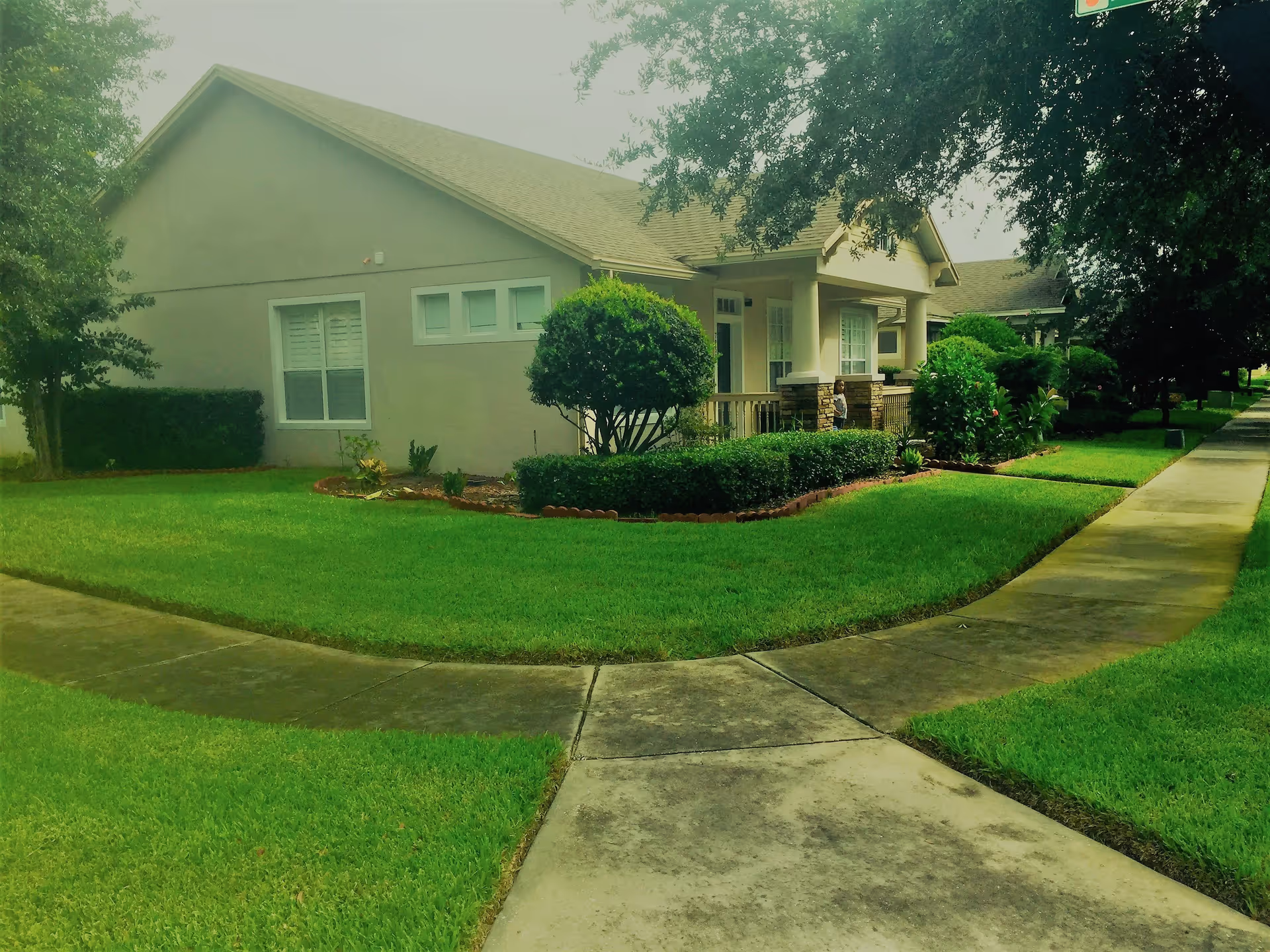 Exterior view of a single-story assisted living facility building with a well-maintained green lawn, bushes, and trees. A concrete sidewalk curves around the front of the building, which has a covered porch area with columns.