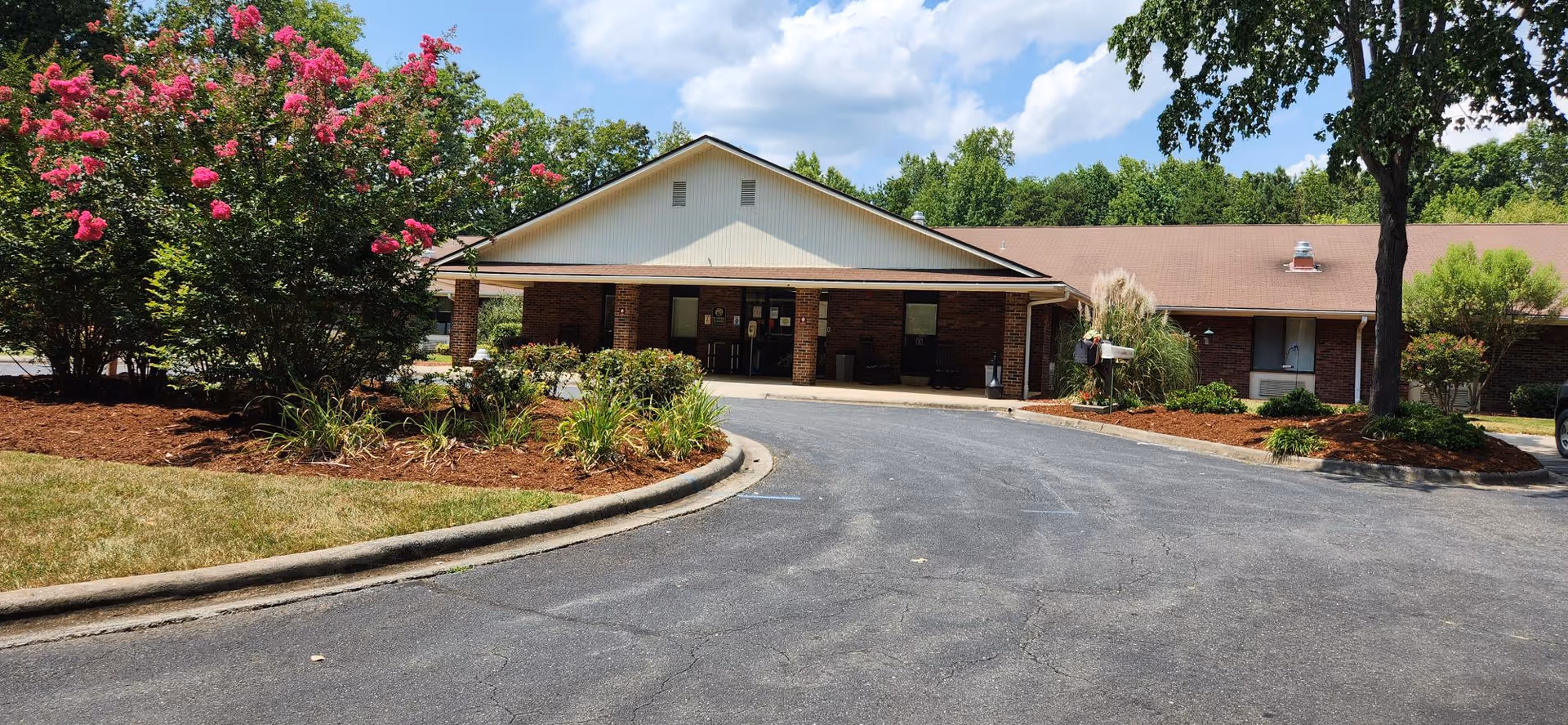 Front exterior view of Asheboro Rehabilitation and Healthcare Center showing a single-story brick building with a covered entrance, surrounded by landscaped greenery and flowering trees under a partly cloudy sky.