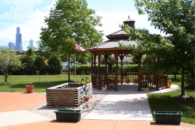 Outdoor garden area with a wooden gazebo surrounded by green trees and grass. There are planters and a small fenced garden bed near the gazebo. A city skyline is visible in the background under a partly cloudy sky.