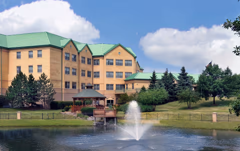Exterior view of Alden Estates of Orland Park, a multi-story building with green roofs, surrounded by trees and landscaping. In the foreground, there is a pond with a water fountain and a wooden gazebo on the edge of the pond.