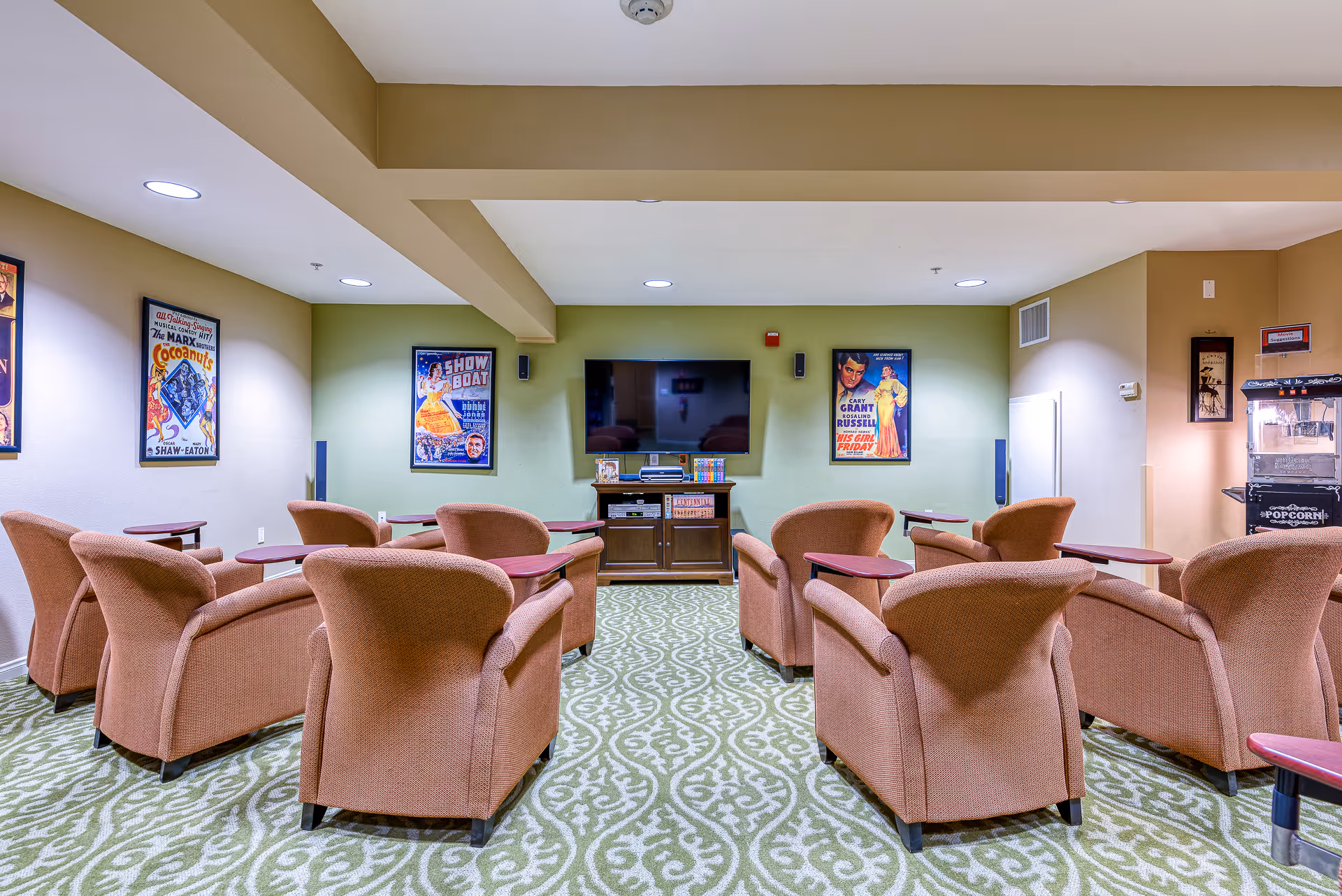 A cozy media room with multiple cushioned armchairs arranged in rows facing a wall-mounted flat-screen TV. The room features a patterned green carpet, framed vintage movie posters on the walls, and a popcorn machine in the corner.