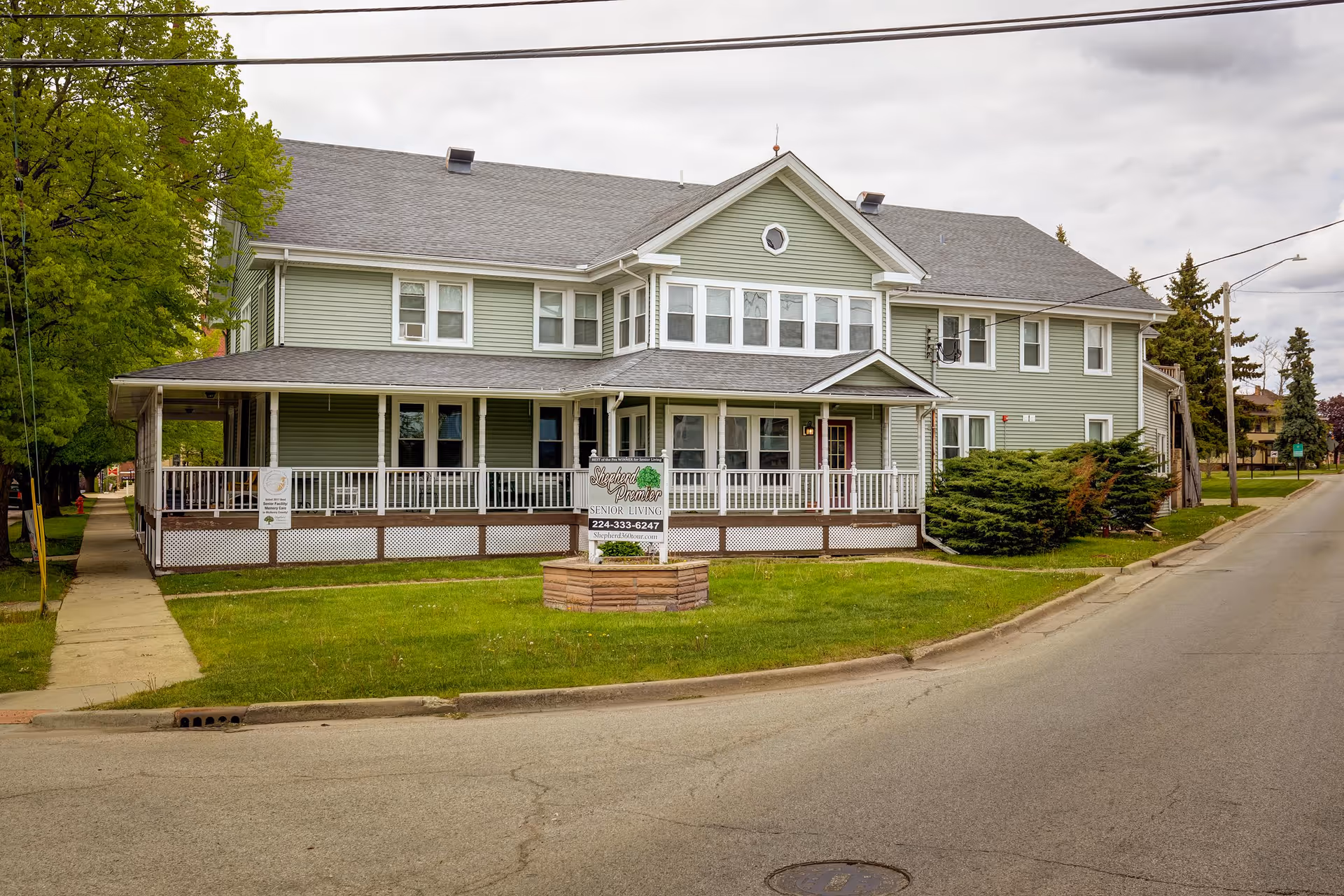 Exterior view of a two-story green senior living facility with a wraparound porch and a sign in front that reads Shepherd Premier Senior Living. The building is situated on a corner lot with a sidewalk and some trees nearby under a cloudy sky.