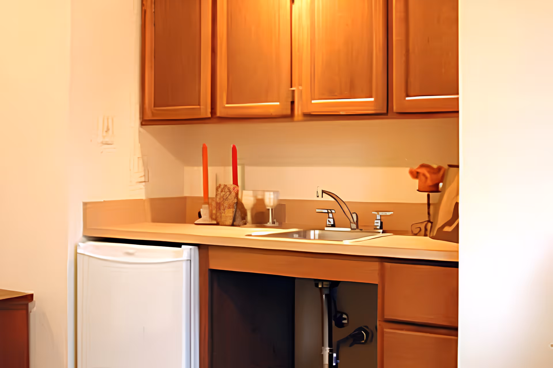 A small kitchen area with wooden cabinets, a countertop with a sink and faucet, two red candles in holders, and a small white refrigerator underneath the counter. The cabinet under the sink is open, revealing plumbing.