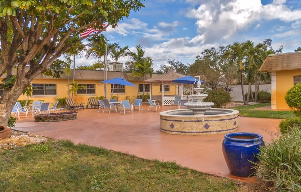 Outdoor patio area at The Residence at Dania Beach featuring a central water fountain, multiple seating arrangements with chairs and tables under blue umbrellas, surrounded by trees and yellow buildings under a partly cloudy sky.