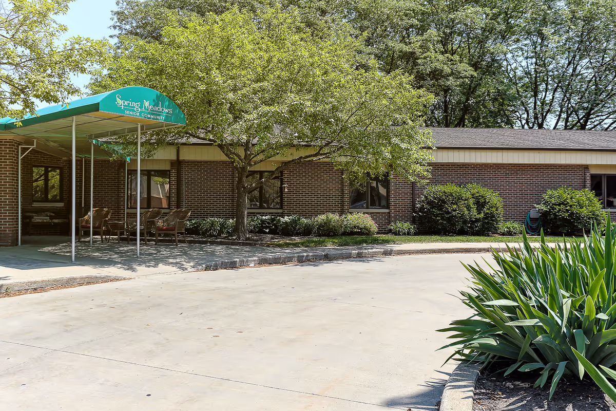Exterior view of a single-story brick building with a green awning that reads 'Spring Meadows Senior Community'. There are trees, shrubs, and a concrete driveway in front of the building.