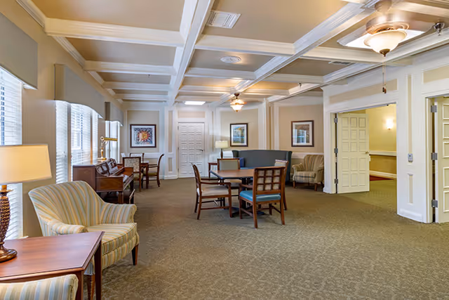 A spacious and well-lit common area with beige walls and carpeted floor. The room features multiple seating arrangements including striped armchairs, wooden chairs around a table, and a small sofa. There is a piano against one wall, framed artwork, and several lamps providing warm lighting. The ceiling has decorative beams and ceiling fans with lights. Large windows allow natural light to enter the room.