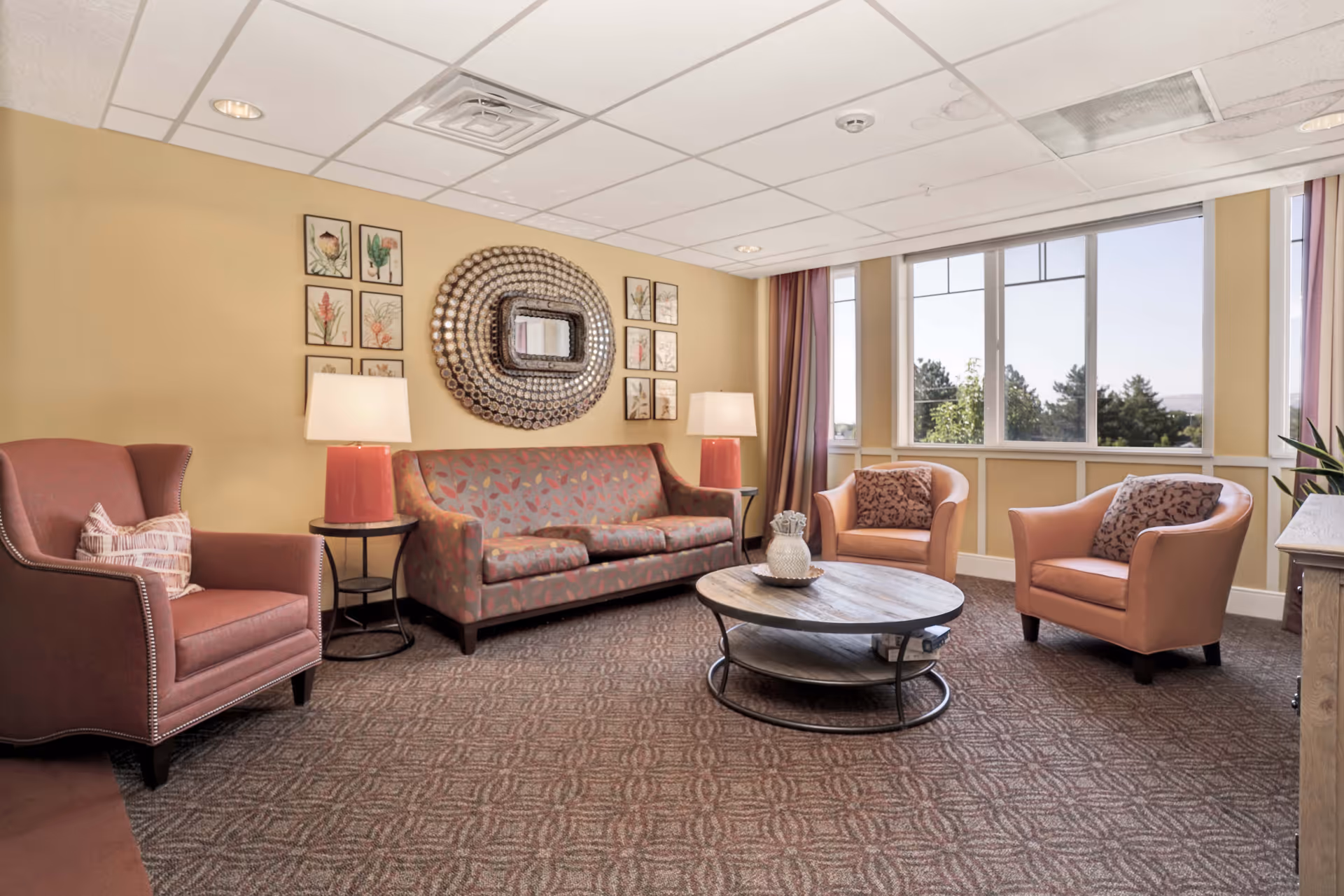 A cozy living room with a patterned sofa, two armchairs, a round wooden coffee table, two side tables with lamps, decorative wall art, and large windows letting in natural light.