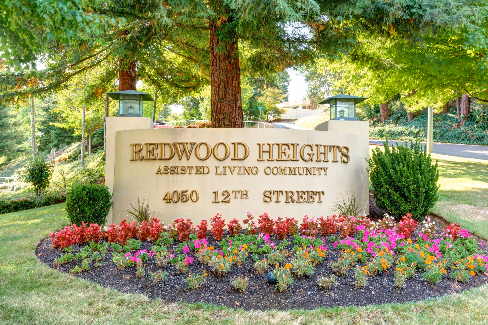 Entrance sign for Redwood Heights Assisted Living Community at 4050 12th Street, surrounded by colorful flowers and greenery with trees in the background.