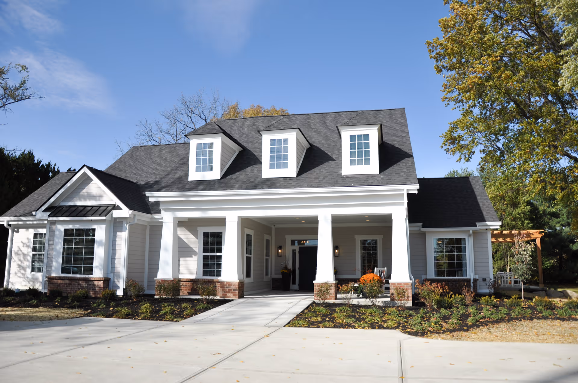 Front exterior view of a single-story residential-style building with white siding, three dormer windows on a dark shingled roof, a covered porch supported by white columns, and landscaped garden beds along the driveway and front walkway.