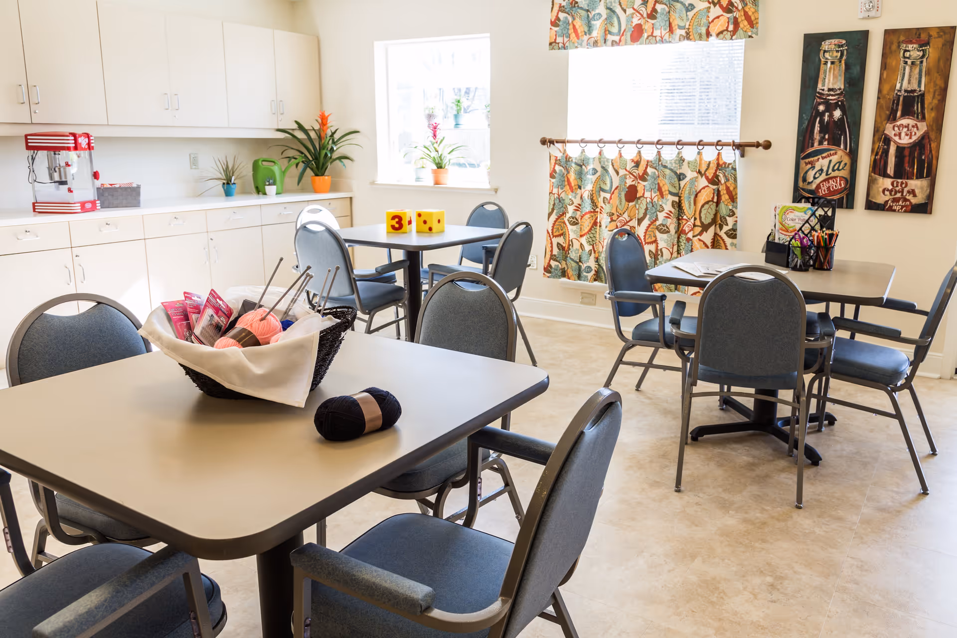 A bright room with several tables and chairs arranged for group activities or dining. On one table, there is a basket containing yarn and knitting needles, and a ball of yarn is placed next to it. The room has white cabinets along one wall, a popcorn machine, potted plants on the windowsill, and colorful curtains with a floral pattern. Two vintage-style soda bottle paintings hang on the wall.