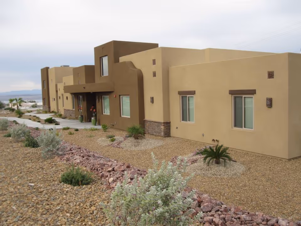 Exterior view of a modern, beige and brown stucco building with multiple windows and a flat roof, surrounded by a desert landscape with gravel, rocks, and sparse vegetation under a cloudy sky.