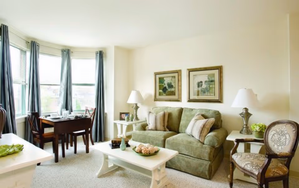 A bright living room with a green sofa adorned with striped pillows, two framed paintings on the wall above the sofa, a white coffee table with decorative items, a side table with a lamp and a small plant, an upholstered armchair, and a dining table with chairs near large windows with blue curtains.