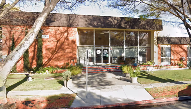 Front exterior view of a single-story brick building with large glass entrance doors, surrounded by trees, plants, and a well-maintained lawn. The building has a sign that reads 'Skilled Care'.