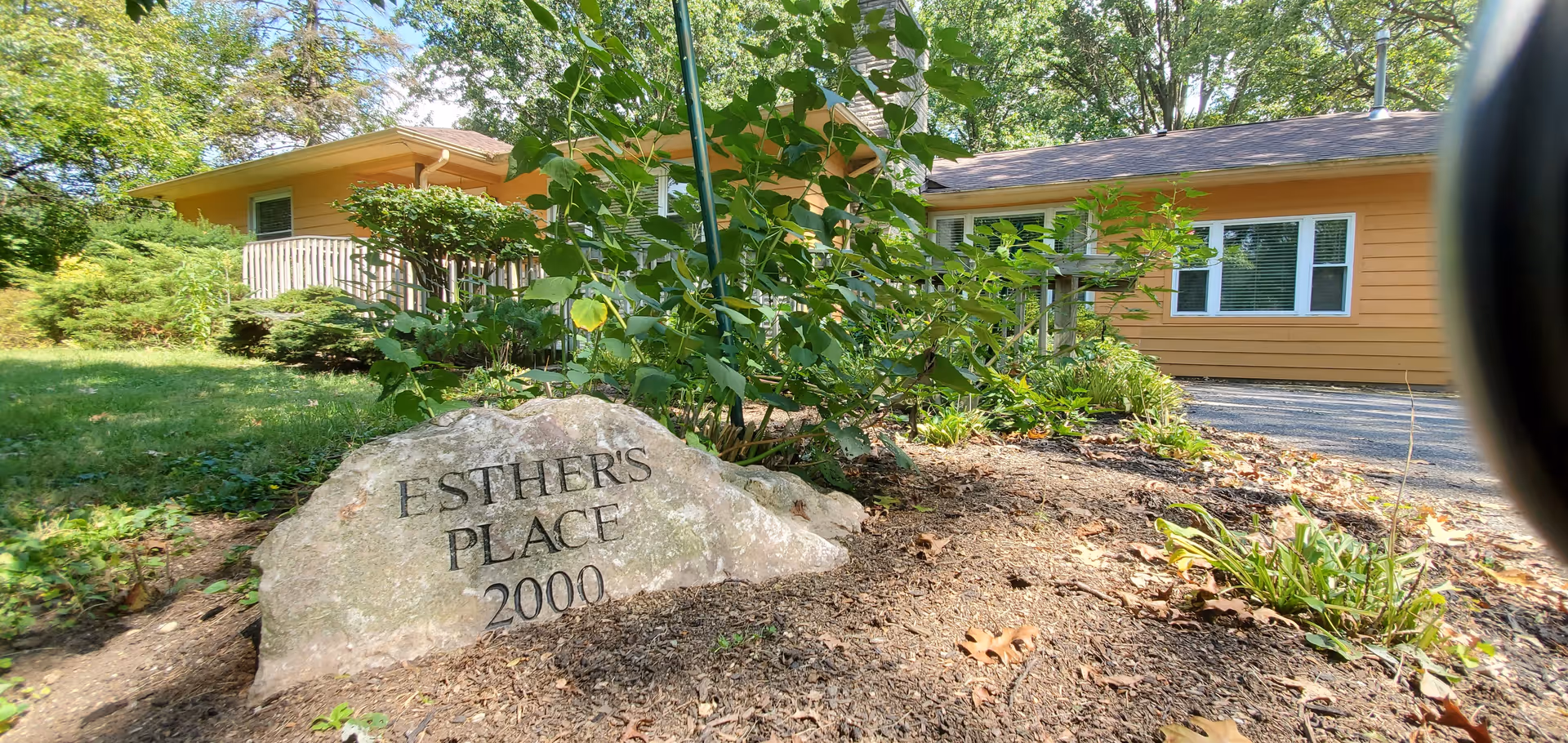 A large rock with the inscription 'ESTHER'S PLACE 2000' in front of a single-story yellow building surrounded by greenery and trees on a sunny day.
