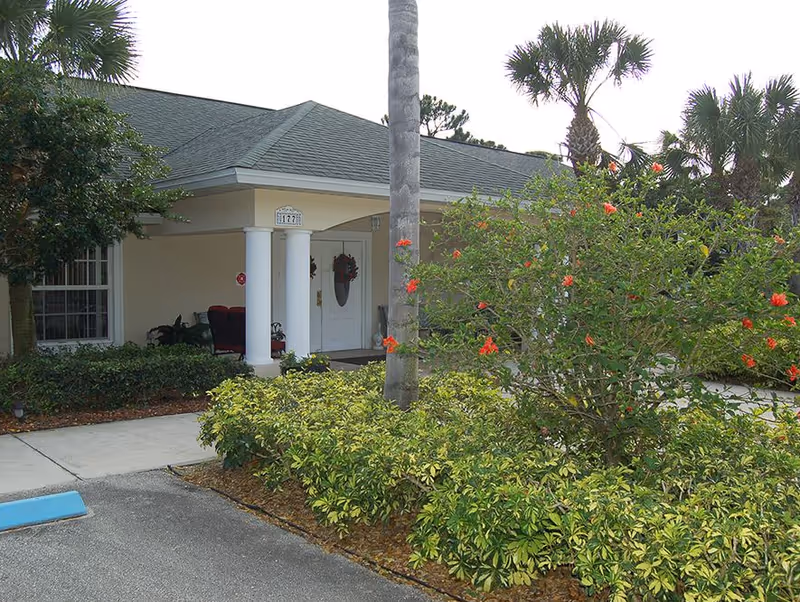 Exterior view of a single-story building entrance with a covered porch supported by white columns. The entrance door has a wreath, and there are chairs on the porch. In front of the building, there are palm trees and flowering bushes with red flowers, along with other greenery. A sidewalk and part of a parking area are visible.