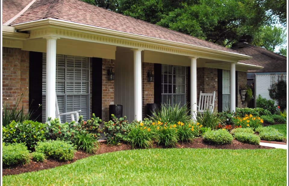 Front porch of a brick building with white columns, black shutters, and white rocking chairs. The porch is surrounded by well-maintained landscaping with green bushes and yellow flowers, and a neatly trimmed lawn in the foreground.