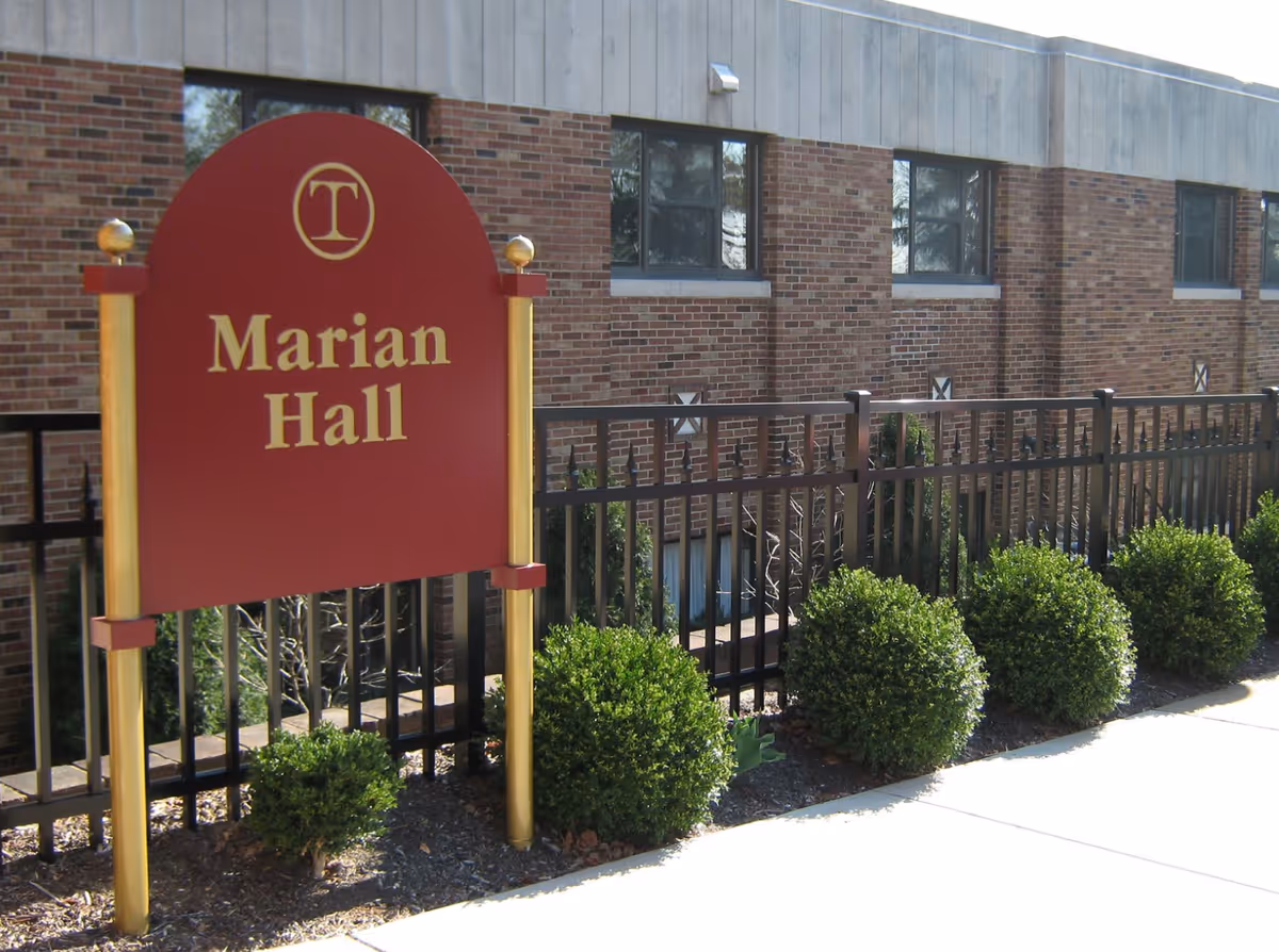 A red and gold sign reading 'Marian Hall' is positioned in front of a brick building with several windows. There is a black metal fence and neatly trimmed green bushes along the sidewalk in front of the building.