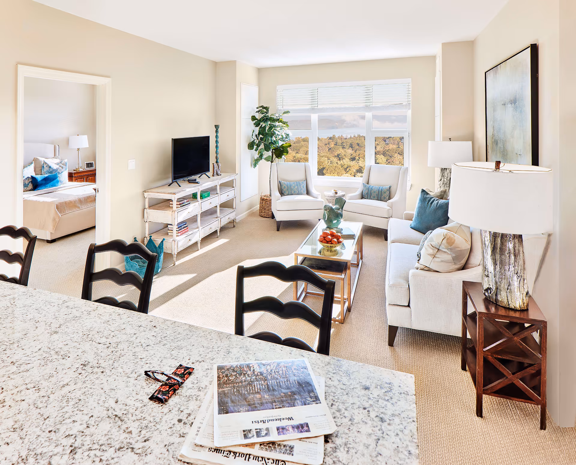 Bright and airy living room with beige walls and carpet, featuring a white sofa with blue and beige pillows, two white armchairs with blue patterned cushions, a glass coffee table with decorative items, a TV on a rustic white console, a large window showing a scenic view of trees, and a glimpse into a bedroom with a bed and nightstand. In the foreground, a granite countertop with newspapers and reading glasses is visible along with three black chairs.